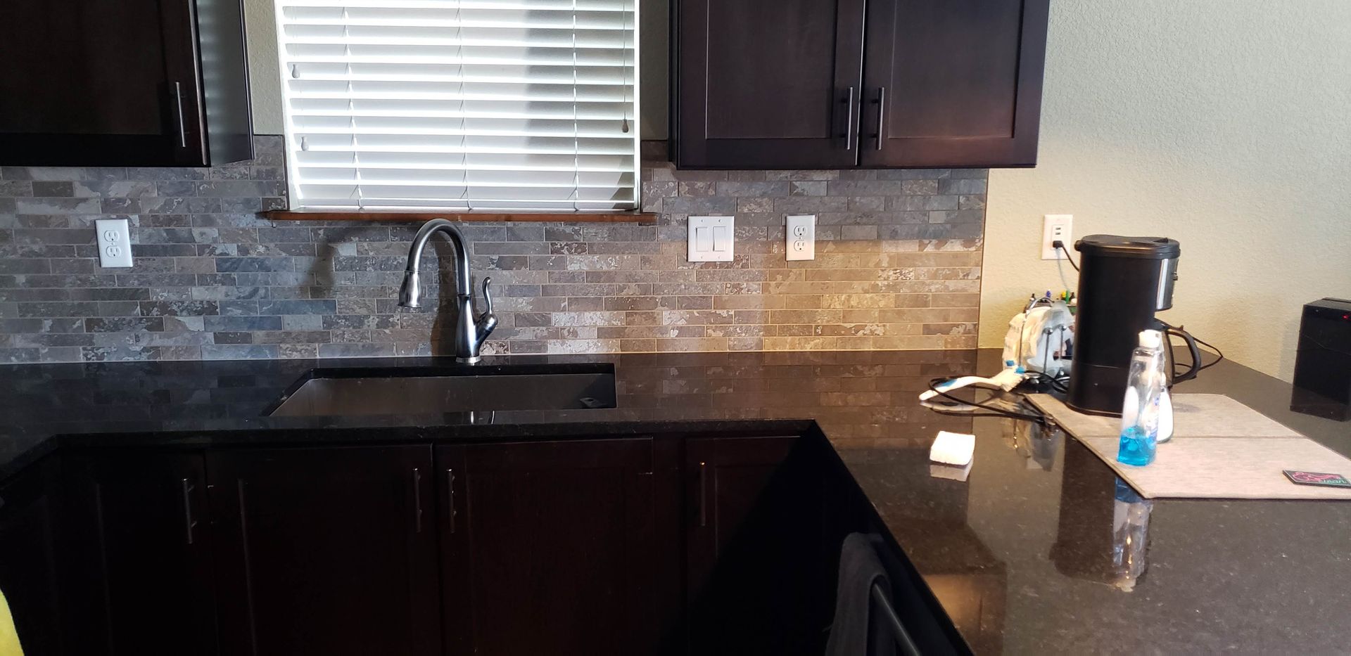 Kitchen countertop and sink with dark cabinets, stone backsplash, window blinds, and a coffee maker on the right.