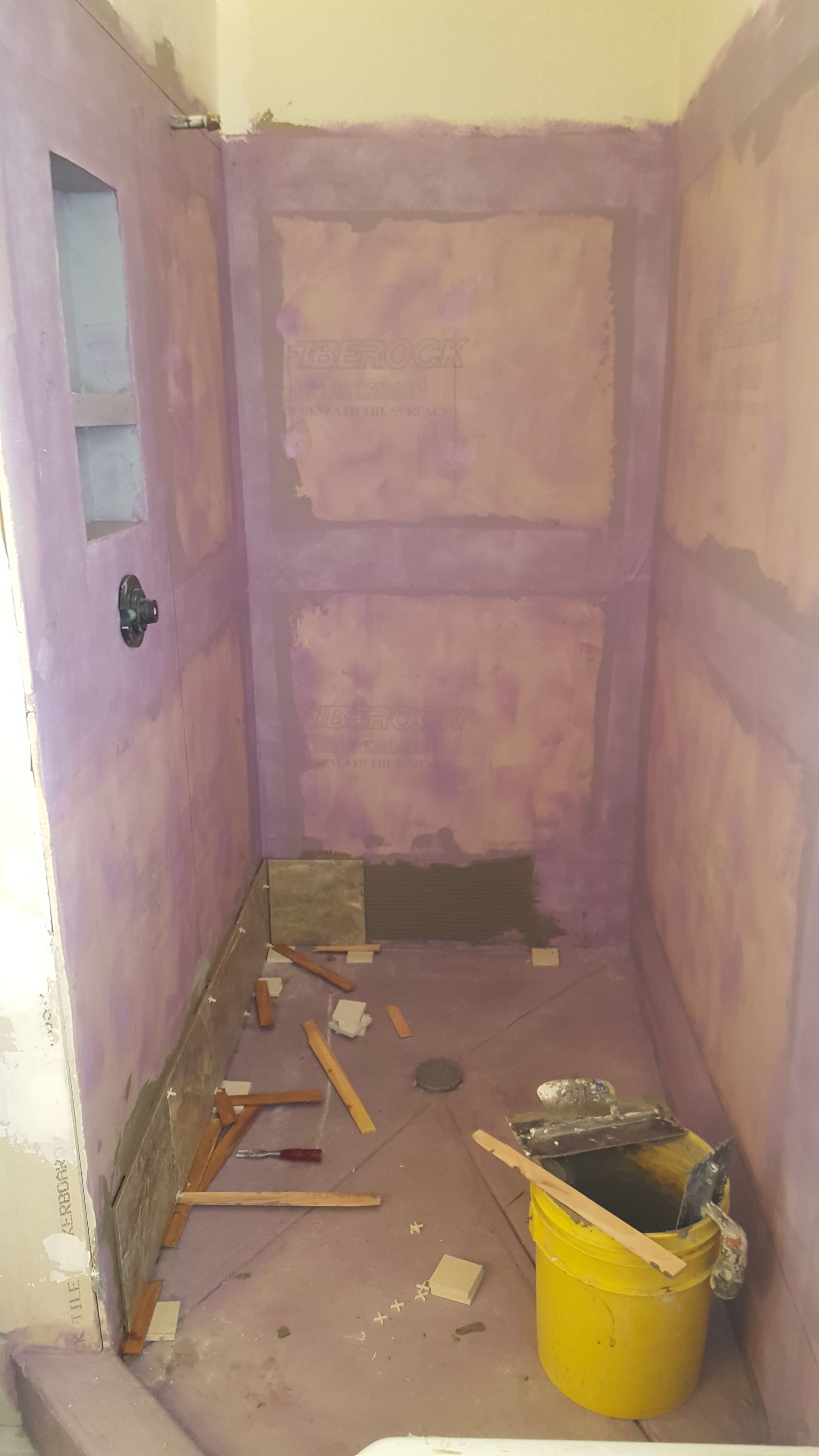 Narrow hallway under renovation with plastered walls, scattered debris, and a yellow bucket on the floor.