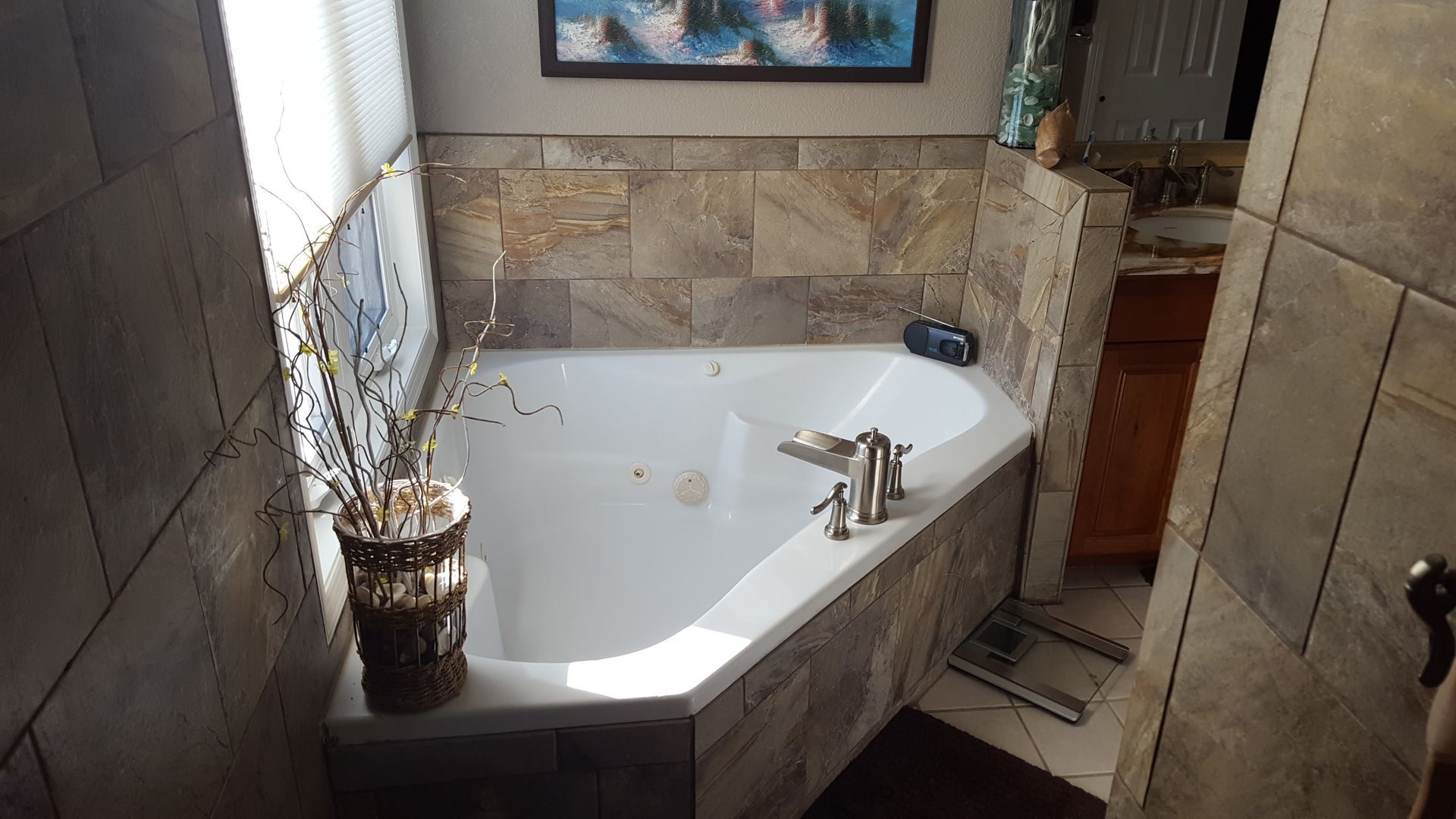 Stone-tiled bathroom with a white corner bathtub and chrome faucet beside a window.