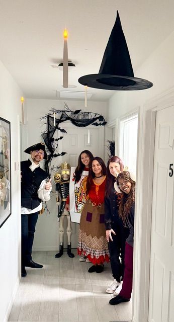 People in Halloween costumes pose in a decorated hallway. Decorations include witch hat, candles, and a skeleton.