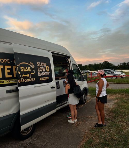 Two women order food from a white food truck, with the sunset in the background.