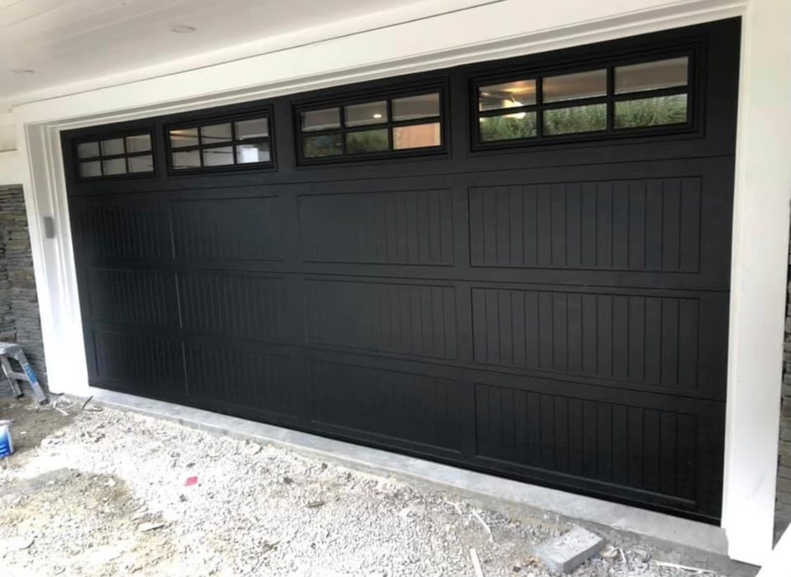 A black garage door with a lot of windows is sitting on top of a gravel driveway.