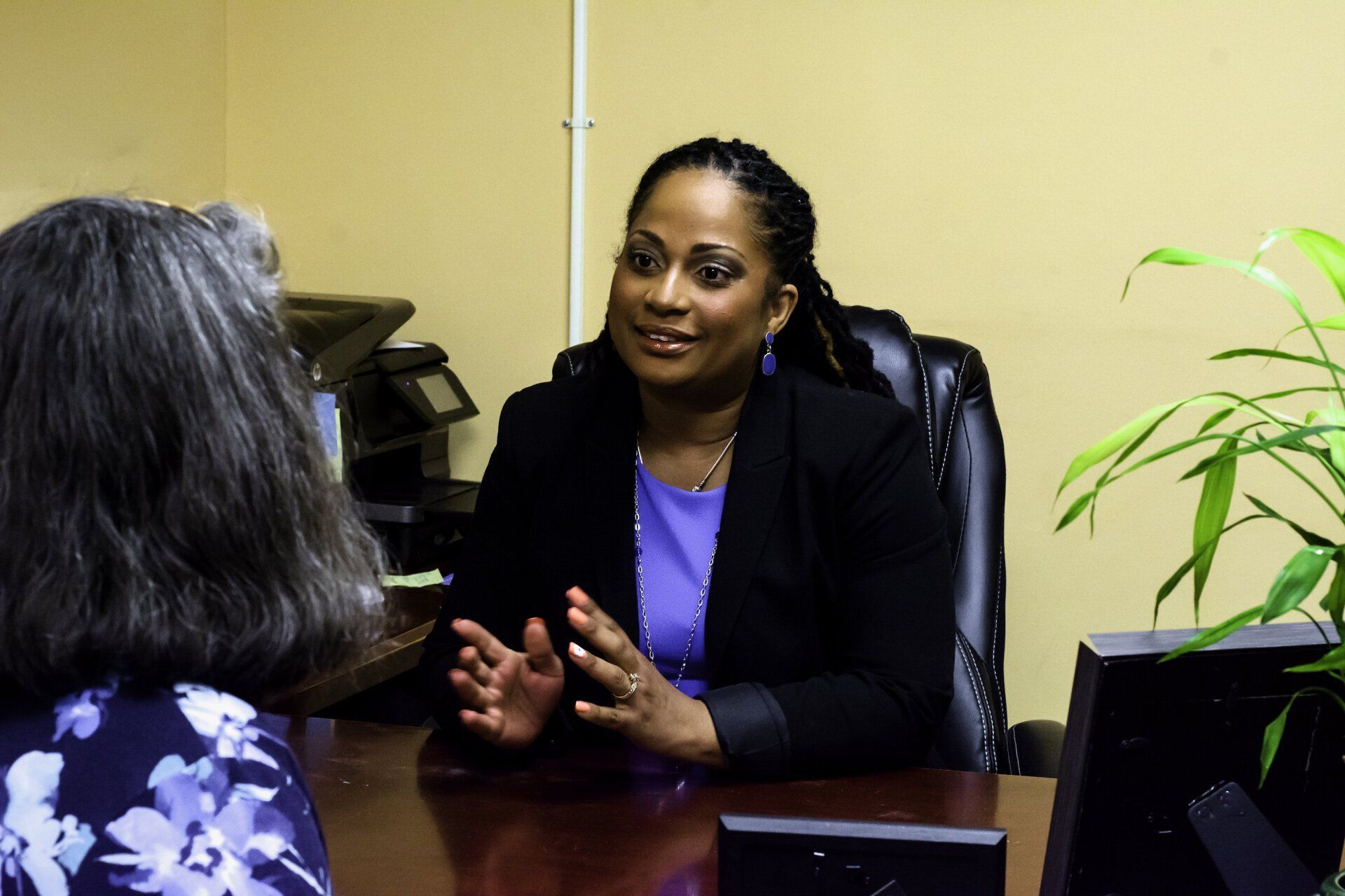 A person in a purple top and black blazer speaks to someone at an office desk, gesturing with their hands.