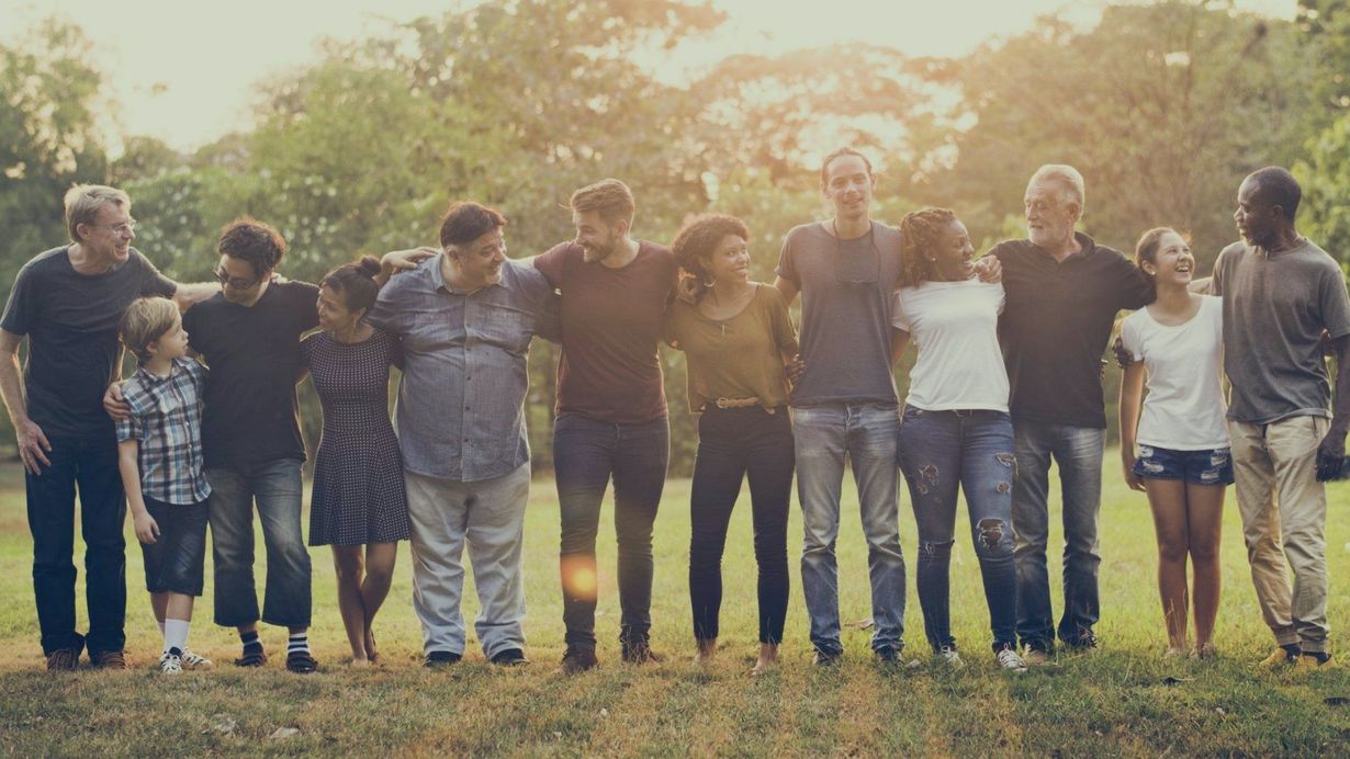 A diverse group of people stand in a line outdoors in a park, arms around each other's waists, smiling together.