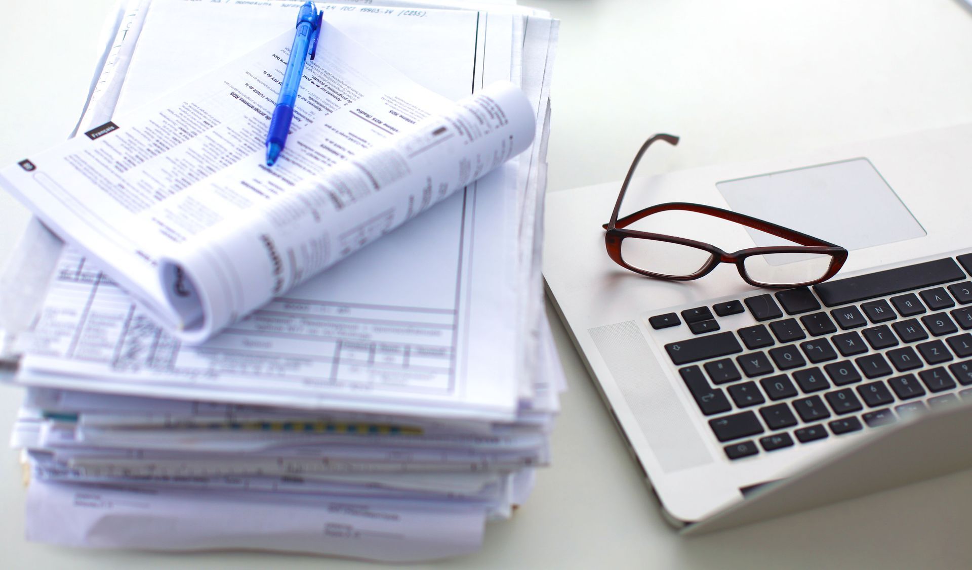 A stack of papers with a pen and glasses next to a laptop.