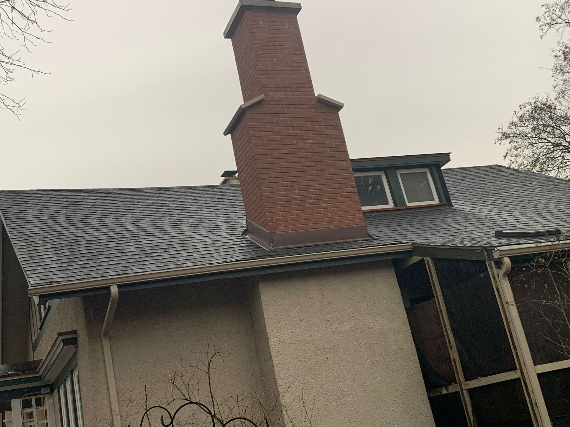 Brick chimney on a house with a dark, wavy roof. Overcast day.