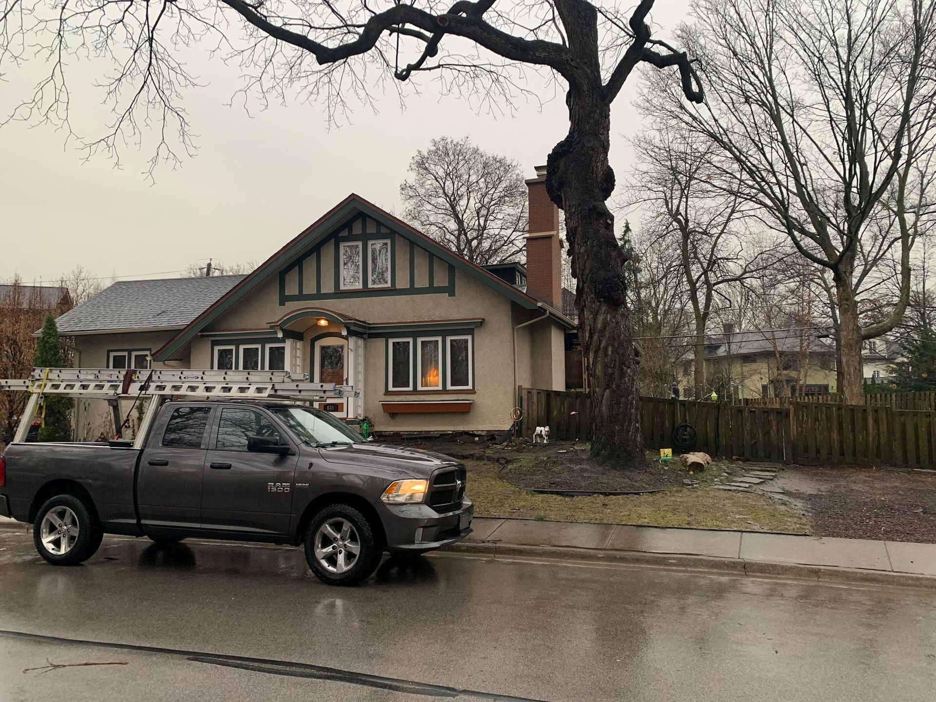 Gray pickup truck parked in front of a house on a wet street. Tree next to the house. Overcast sky.