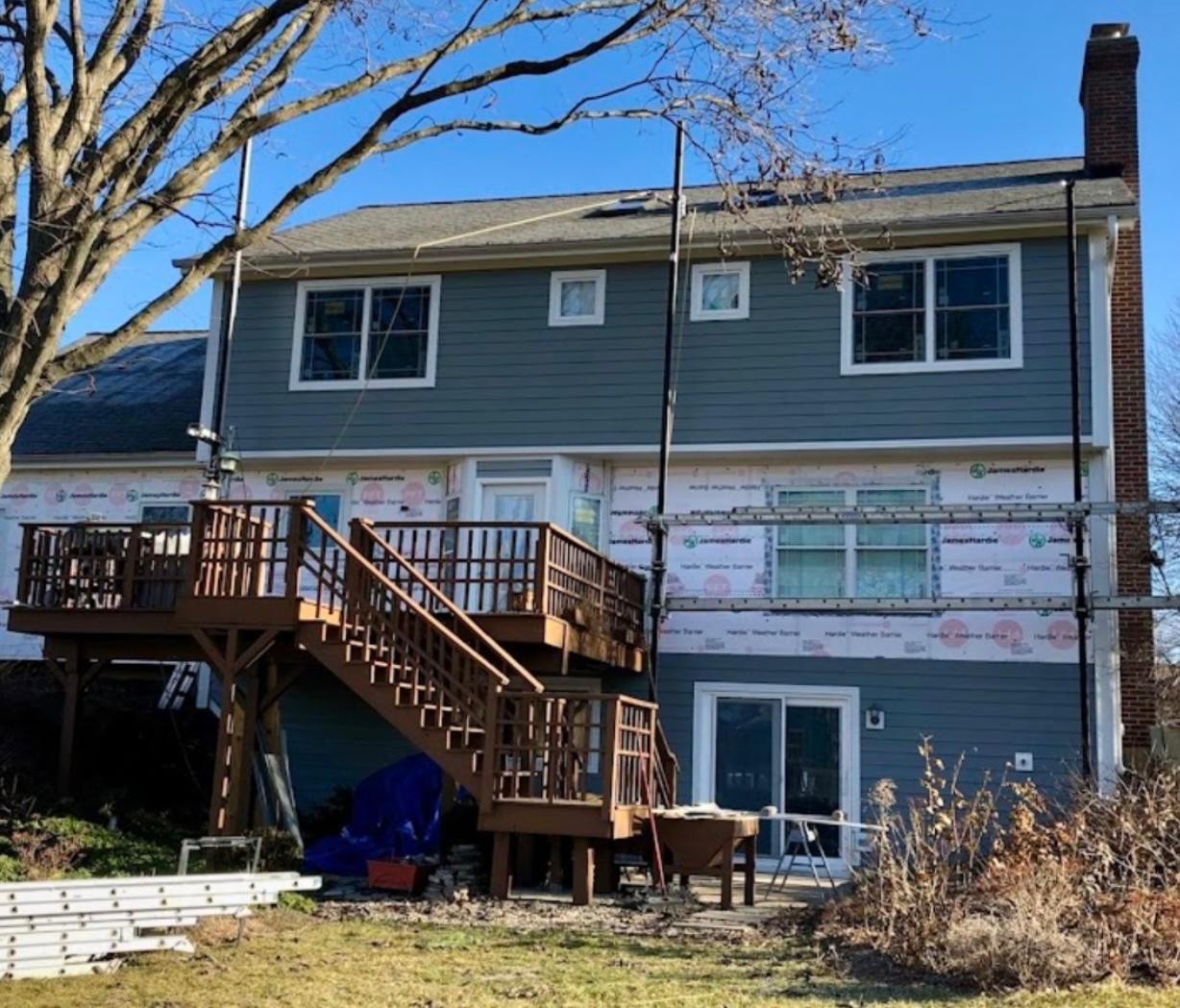 Back of a two-story house under construction; blue siding, wooden deck, scaffolding, and a brick chimney.