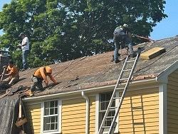 Roofers removing old shingles from a yellow house on a sunny day.