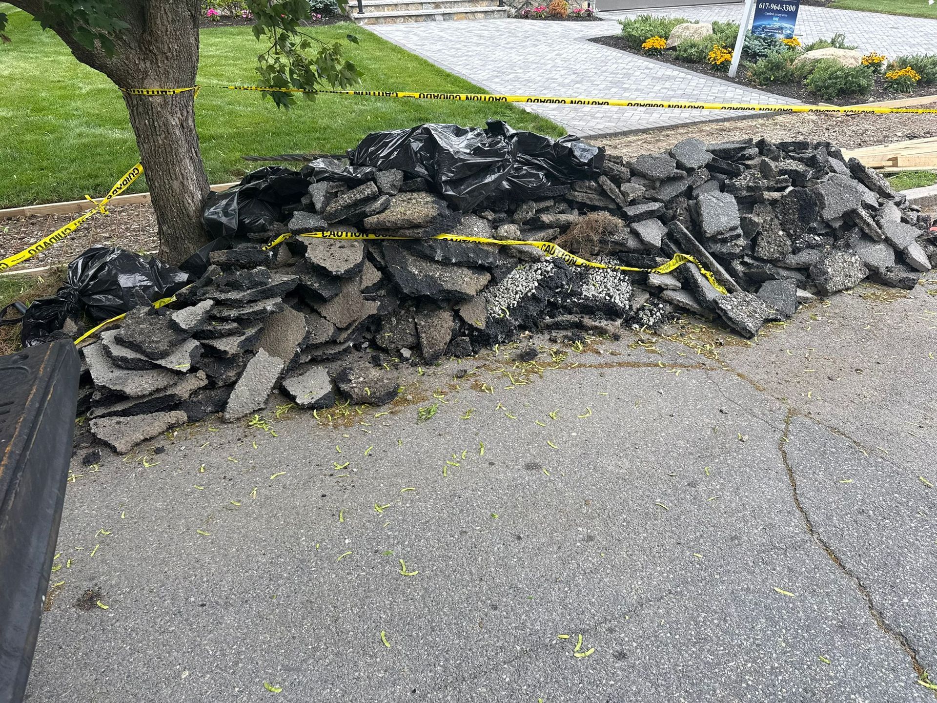 Pile of broken asphalt on a street next to a tree, with yellow caution tape visible.