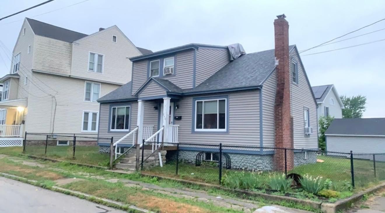 Gray house with a brick chimney, a small front yard with a black fence, and other houses in the background.