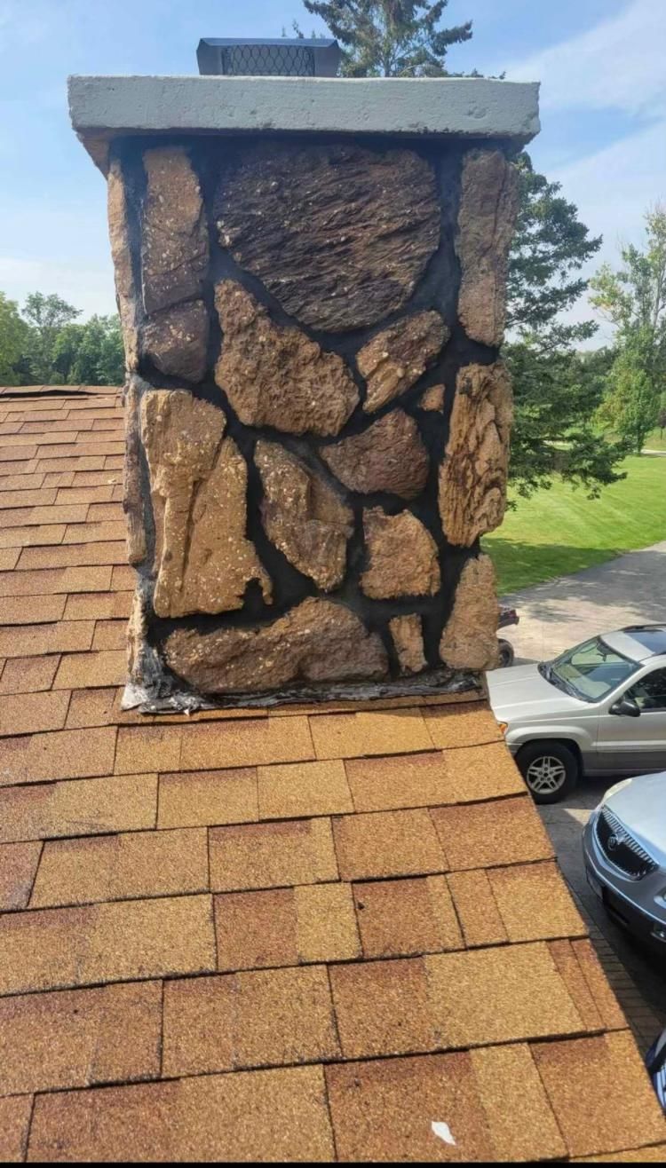 Chimney made of stone on a brown shingled roof, topped with concrete. A car is parked nearby.