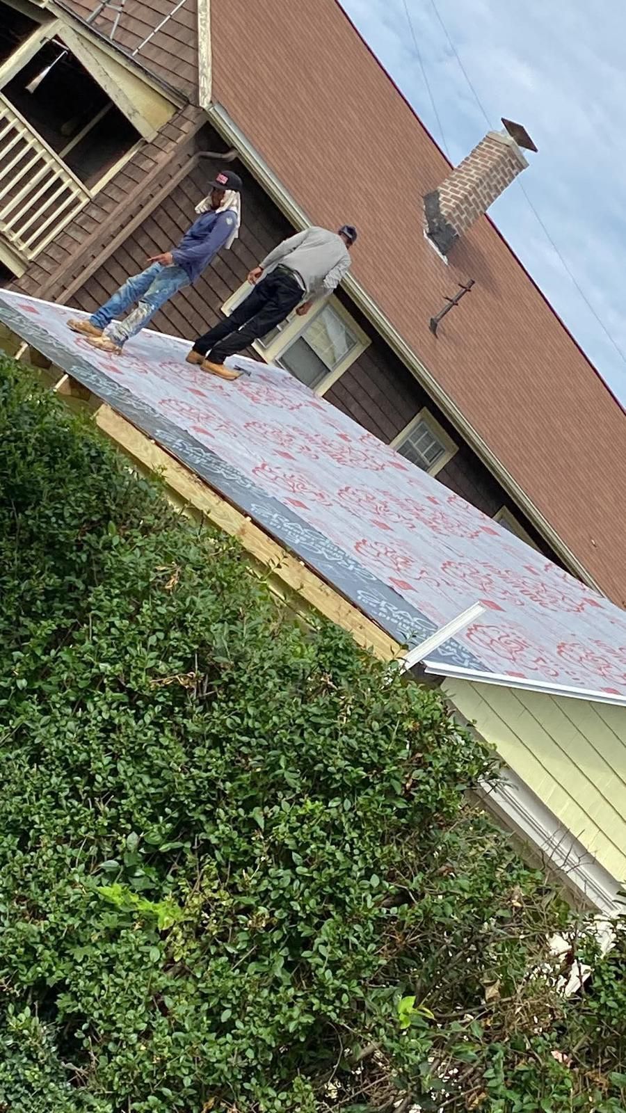 Two people stand on a house roof covered in debris. One wears a blue shirt, the other a gray jacket.