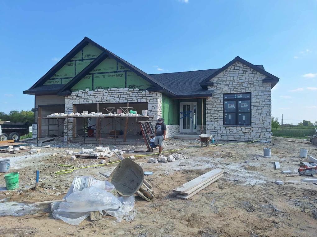 House under construction with stone facade, scaffolding, workers, and building materials.