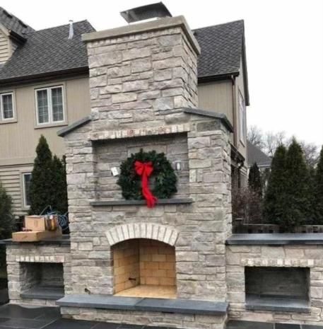 Stone outdoor fireplace with wreath and storage. Brick chimney, tan house in background.