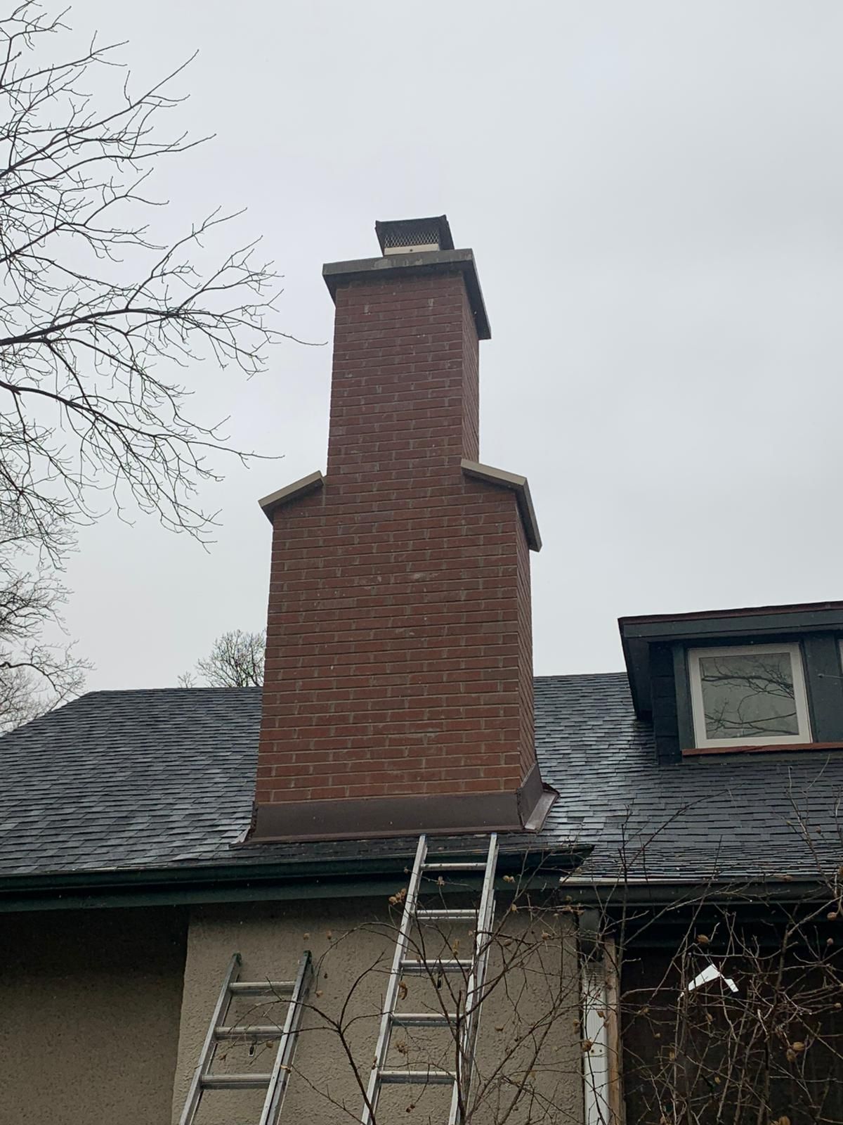 Brick chimney on a roof with a ladder leaning against it, under an overcast sky.