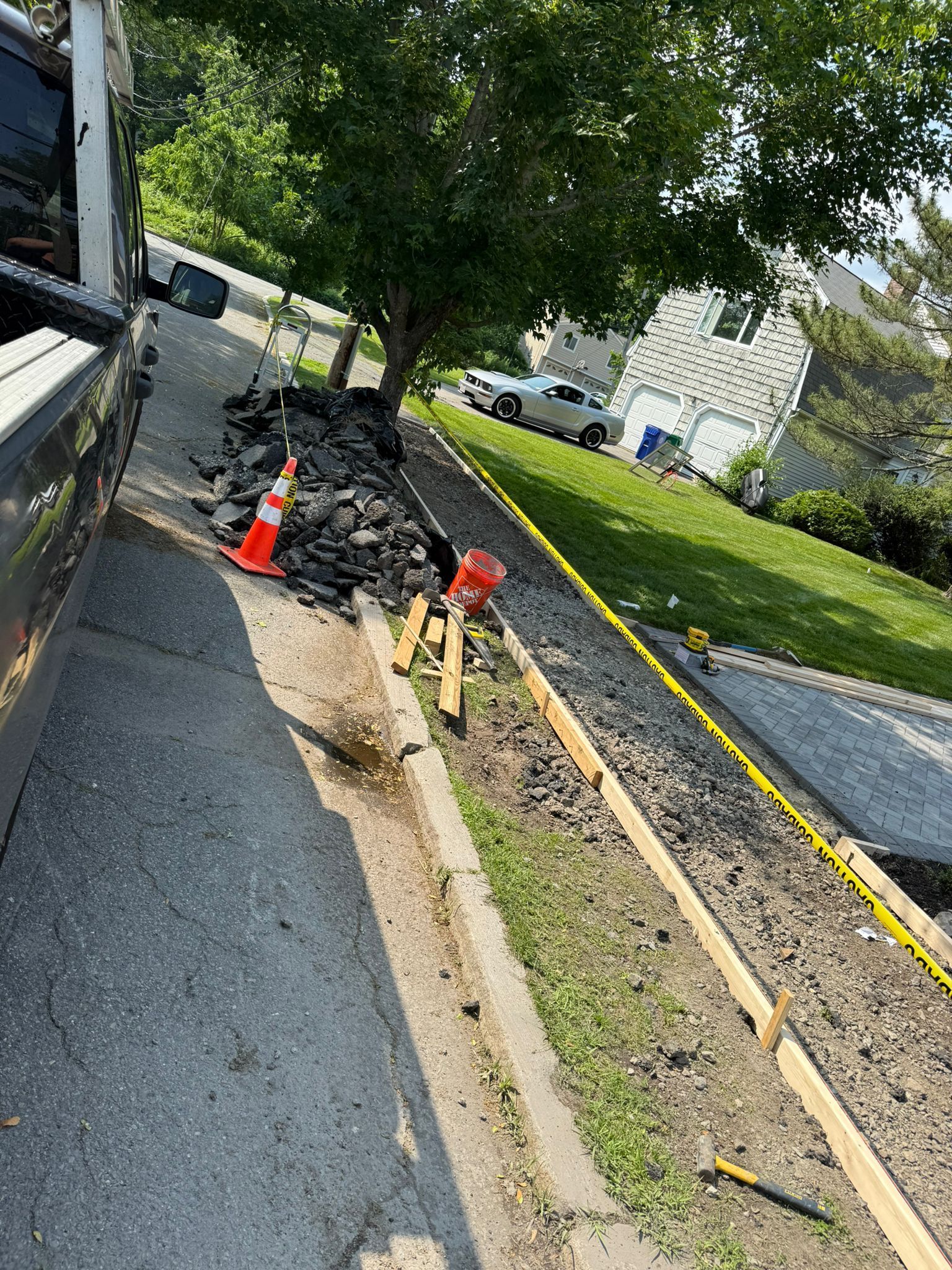 Asphalt driveway with construction. Concrete forms along a grassy strip, rock pile, and an orange cone.