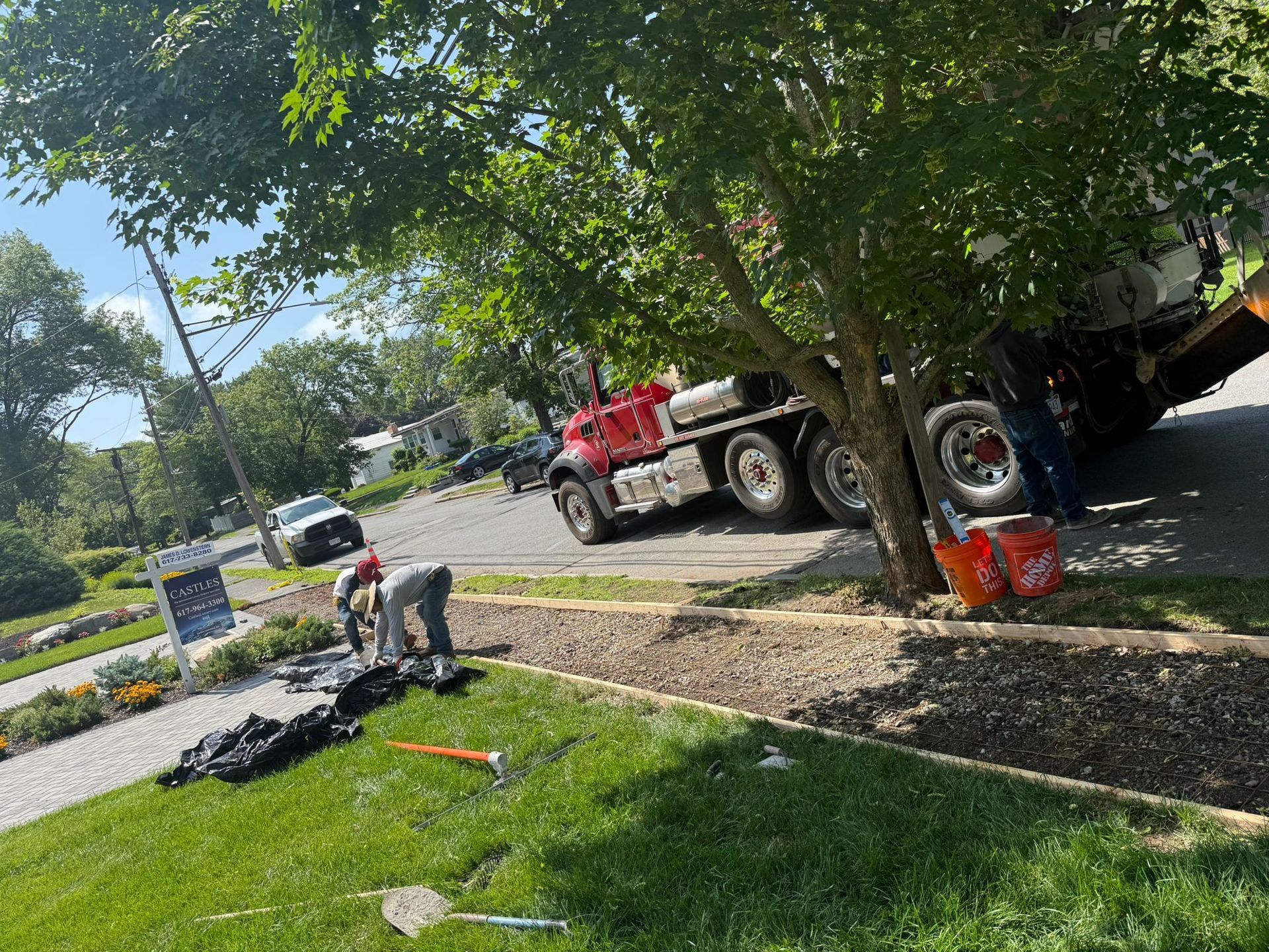 Workers install landscaping next to a residential street with a large truck. Green grass and trees frame the scene.