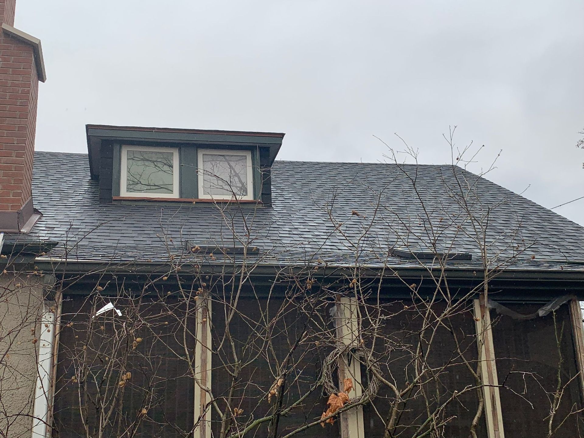 A weathered house roof with a dormer window, visible against a cloudy sky. Bare branches obscure the facade.