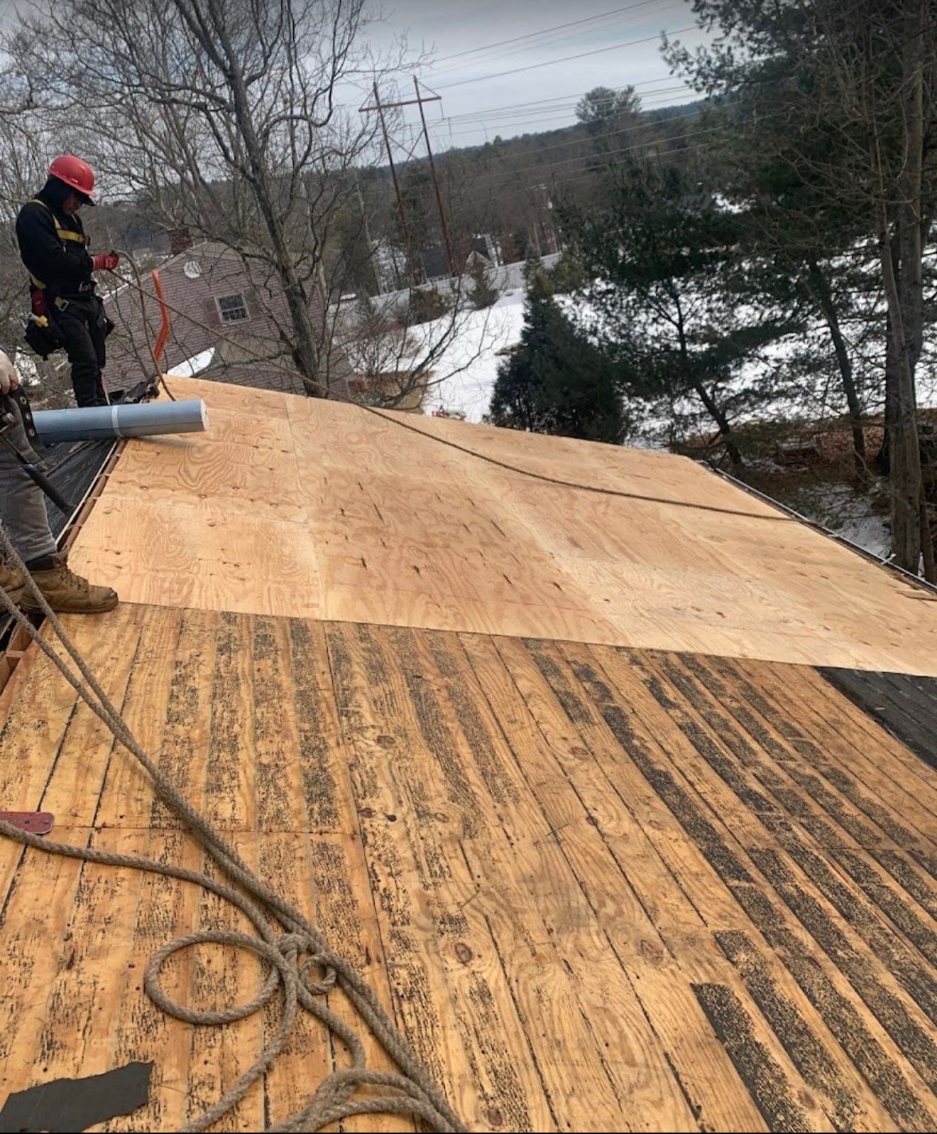 Roofer on a partially re-sheeted roof, wearing safety gear. Winter landscape with snow in the background.