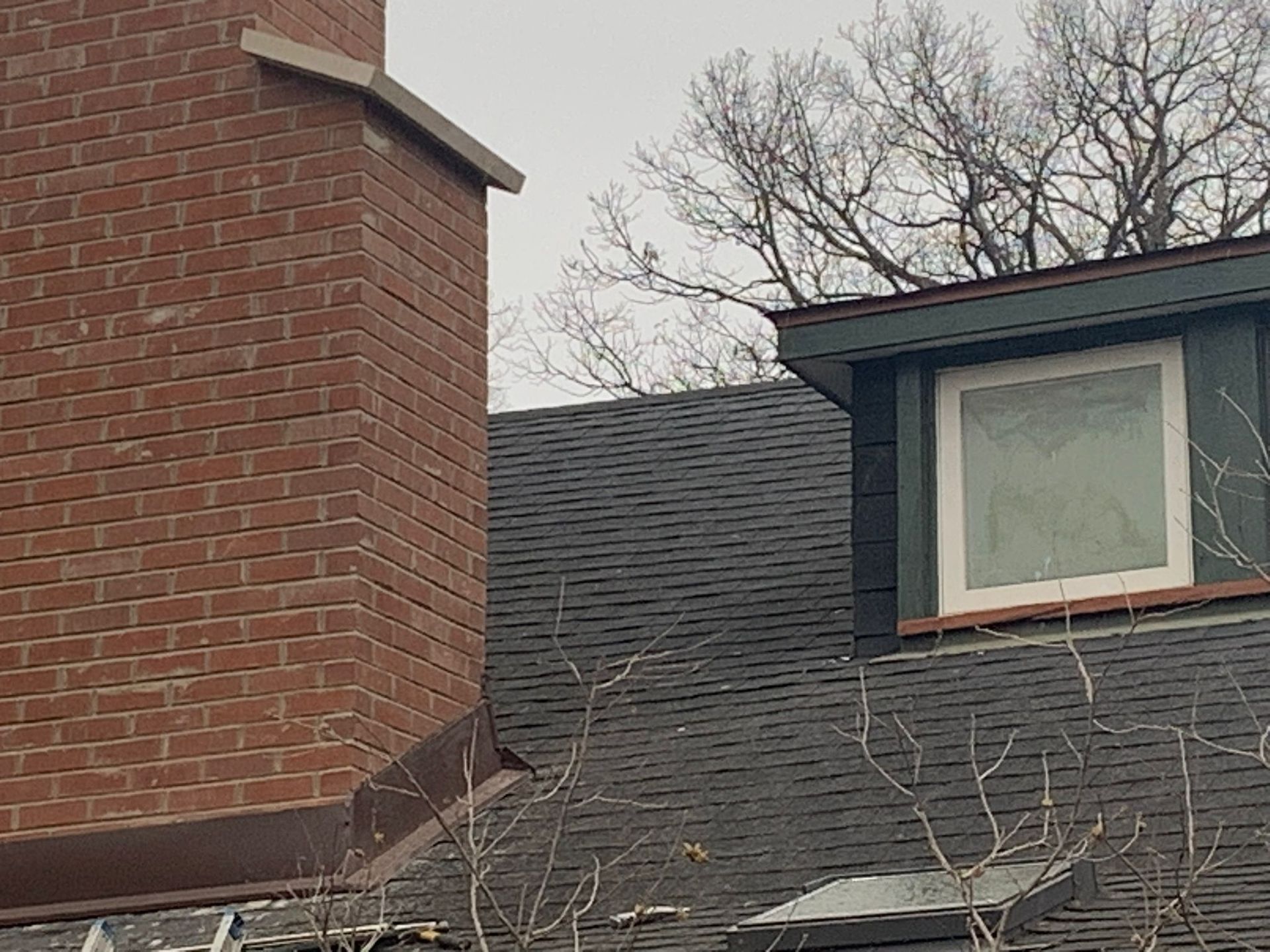 Brick chimney and shingled roof with a dormer window. Bare trees against a cloudy sky.