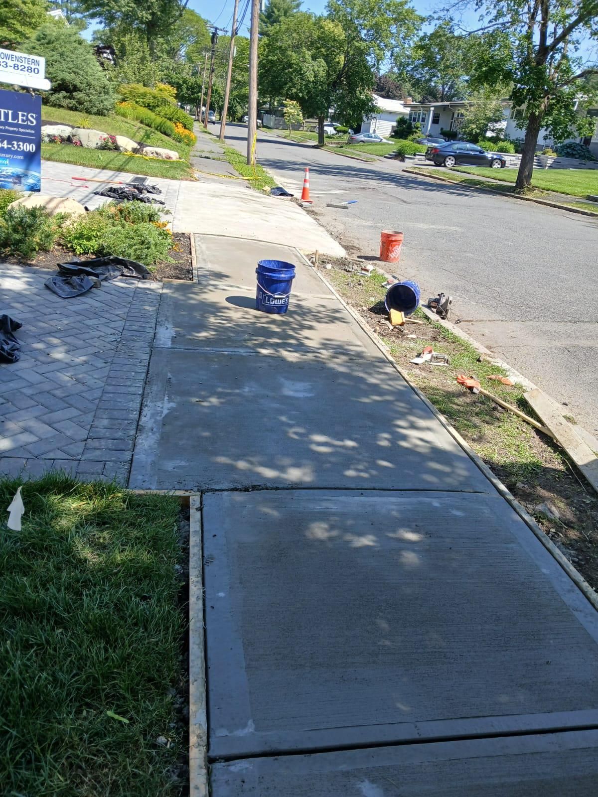 Newly poured concrete sidewalk with a section of brick pavers. Buckets and tools sit nearby on a sunny street.