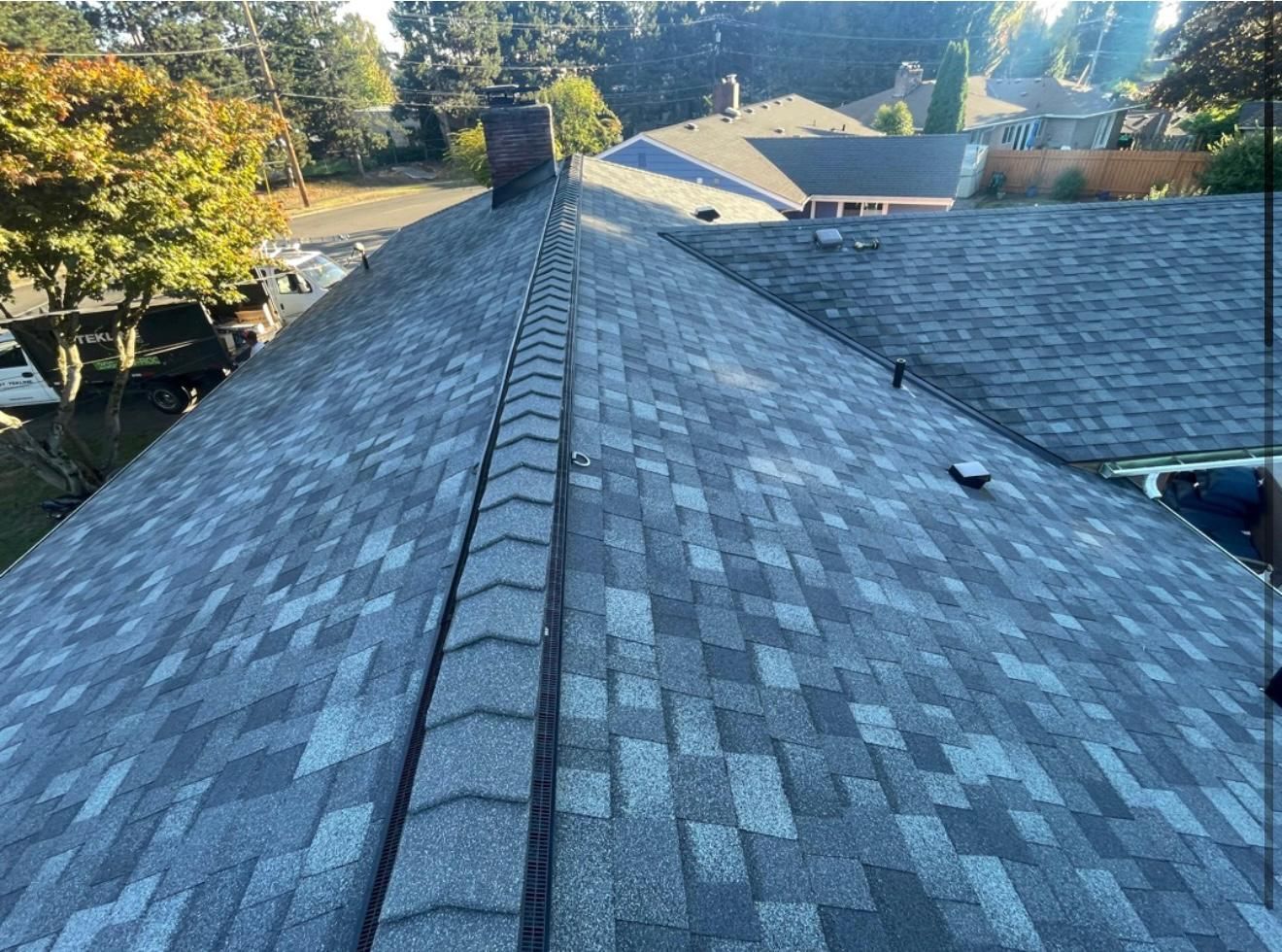 View of a shingled roof with a central ridge and chimney, in an outdoor setting.