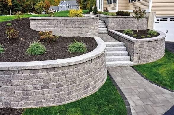Stone retaining walls and stairs leading to a house, with landscaped beds and a paved walkway.