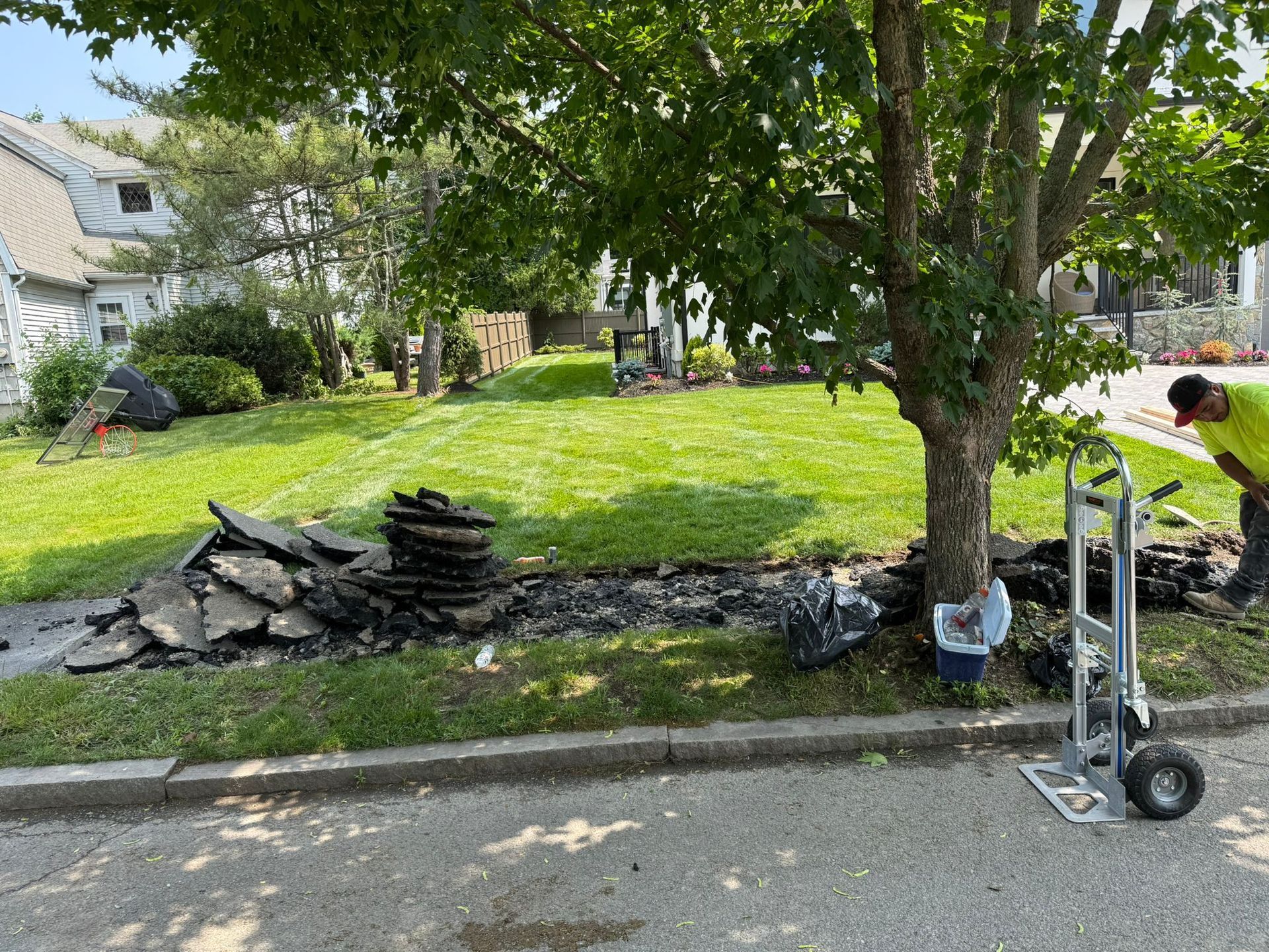 Workers removing asphalt from a yard near a tree, with a dolly, house in the background. Sunny day.