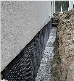 Foundation drainage system installed against a stucco wall: black dimpled membrane, gravel, and excavated earth.