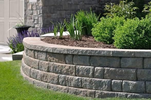 Curved retaining wall made of gray stone blocks with a garden of green plants.