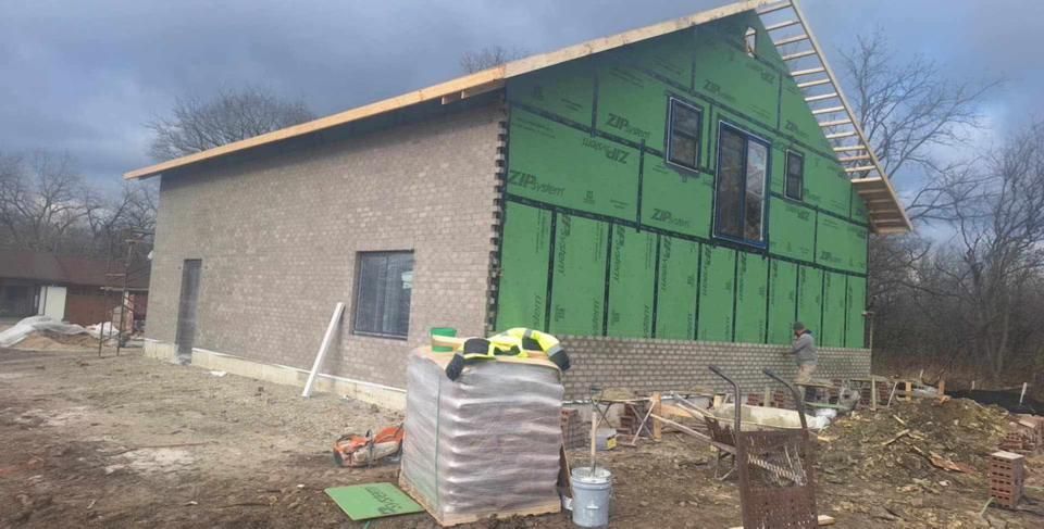 Building under construction, partially bricked, green sheathing, unfinished roof. Supplies and tools visible.