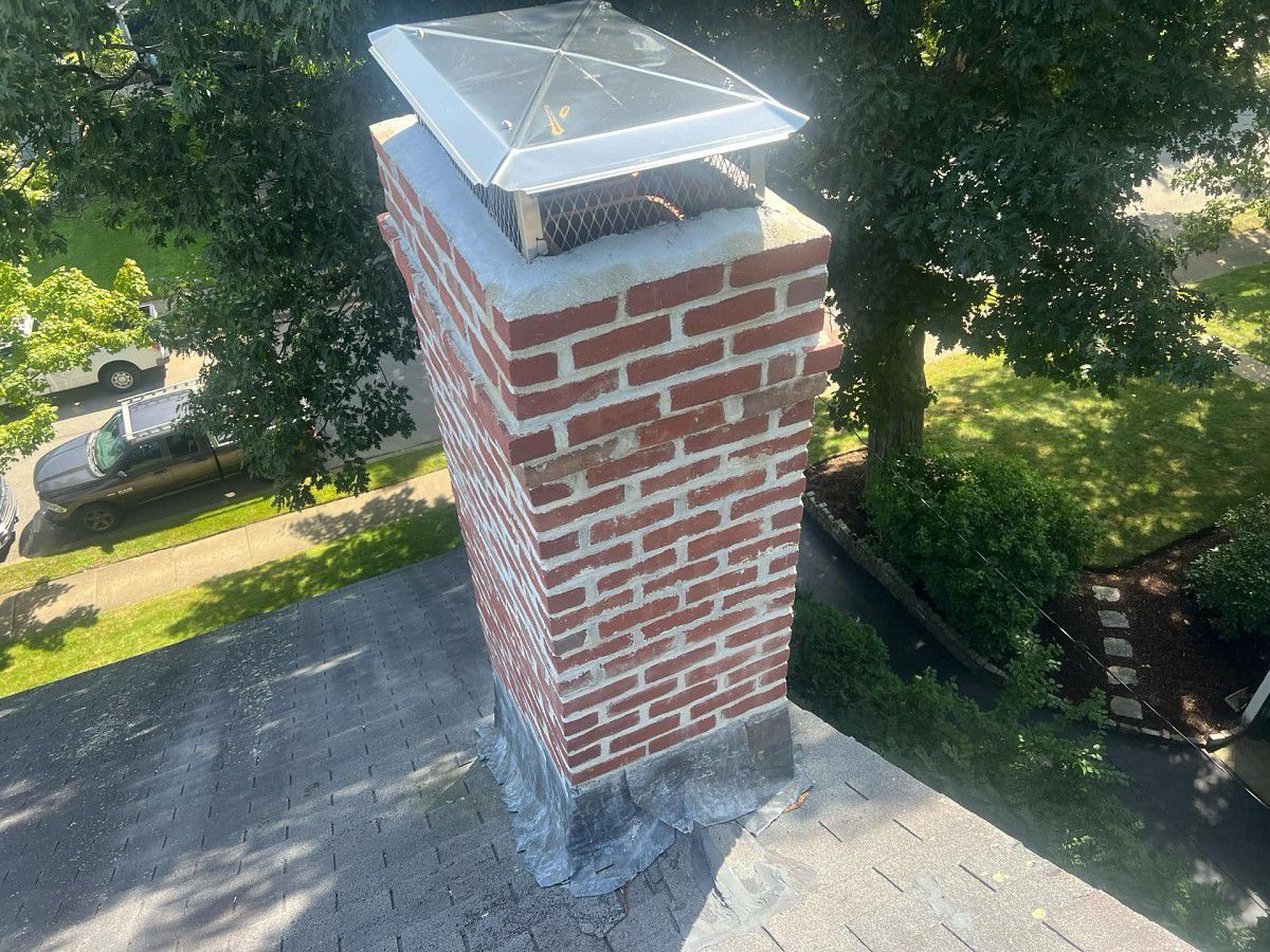 Brick chimney on a gray shingled roof, topped with a metal cap, trees in background.