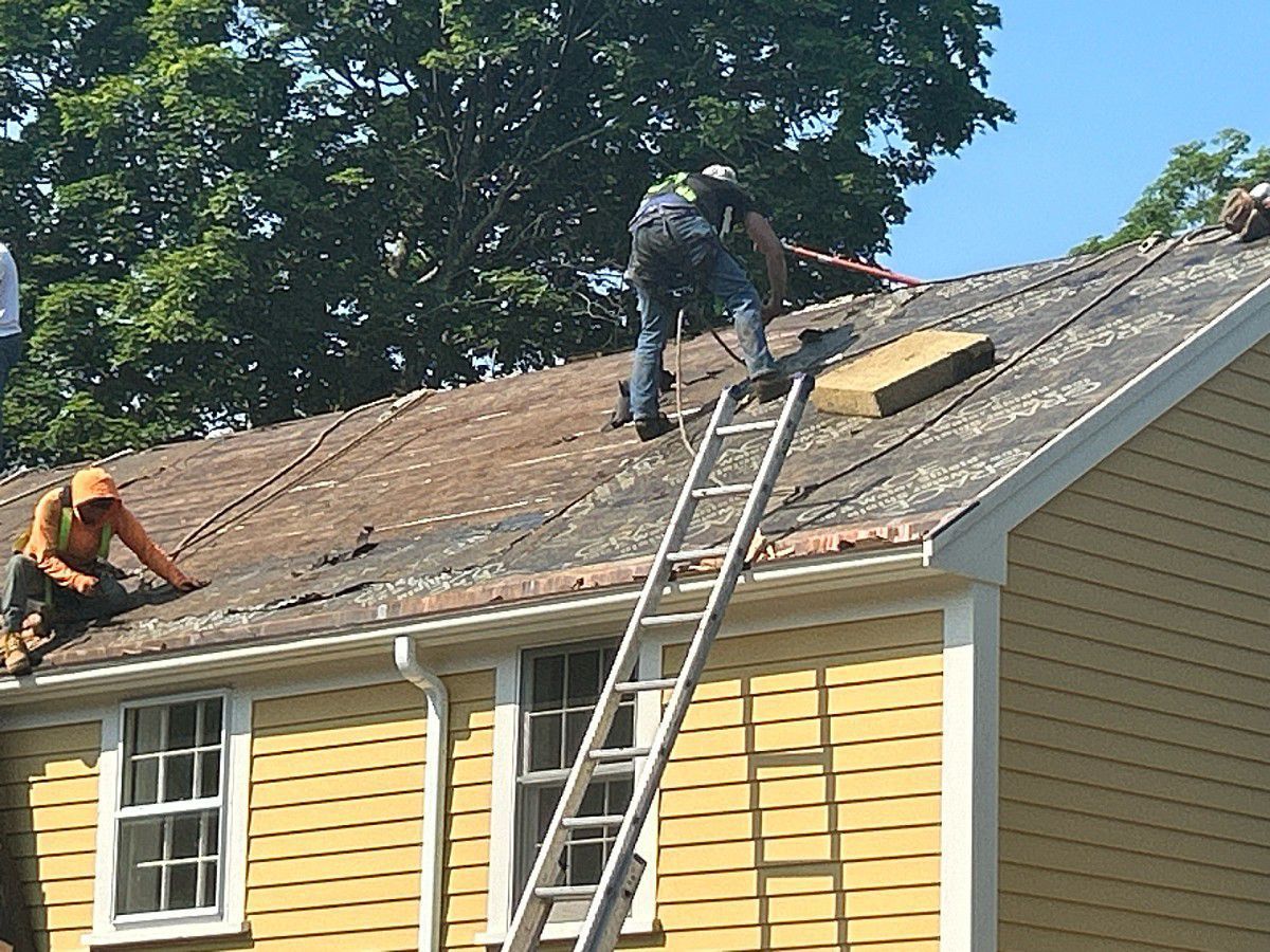 Workers on a yellow house roof, removing shingles. One on a ladder, another crouched. Blue sky, sunny day.