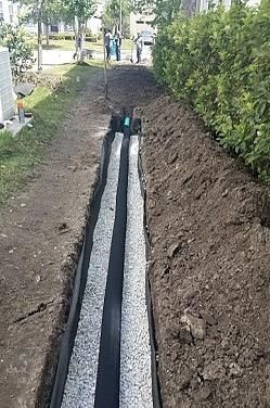 Construction of a trench drain with gravel and dark lining in a grassy area next to bushes.
