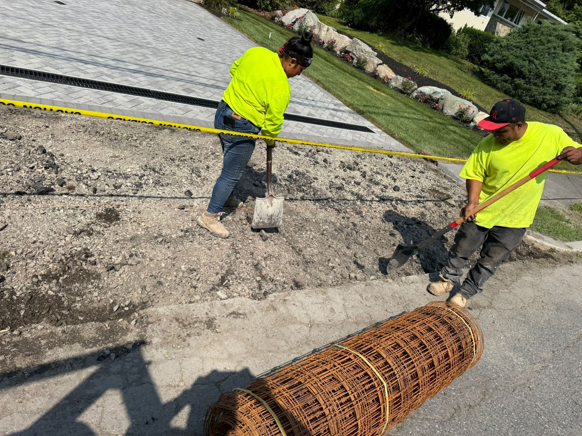 Two people in safety vests working on a hillside; one with a shovel, the other with a rake. A roll of wire mesh lies in the foreground.
