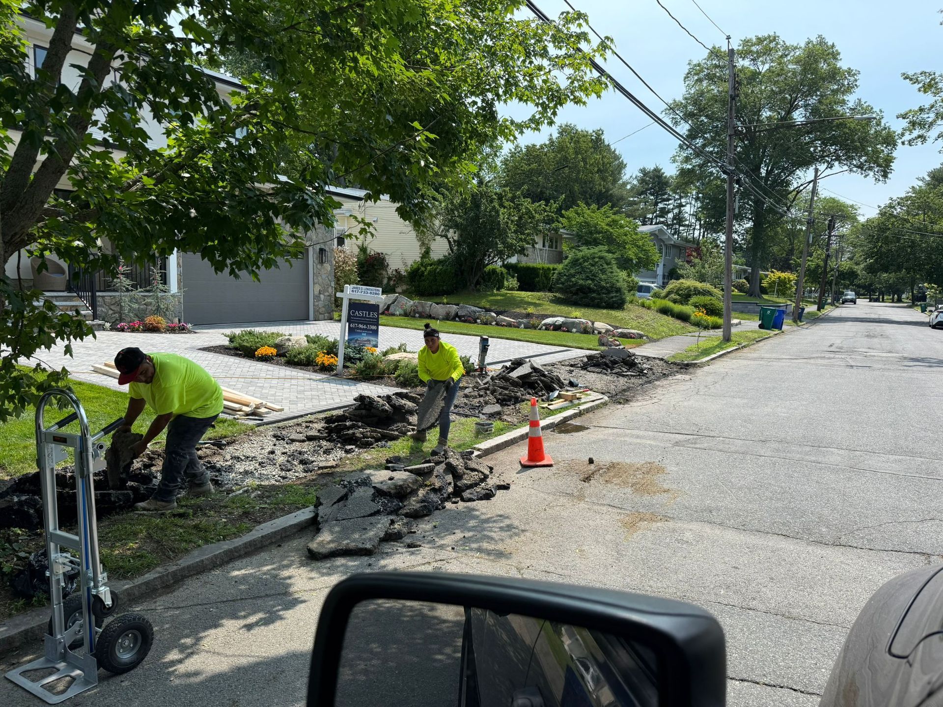 Two workers in neon vests repairing a road, one using a dolly. Asphalt debris, sidewalk, and street present.