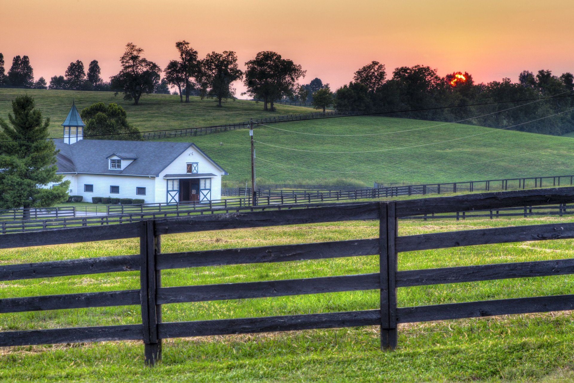 A barn is behind a wooden fence in a field at sunset.