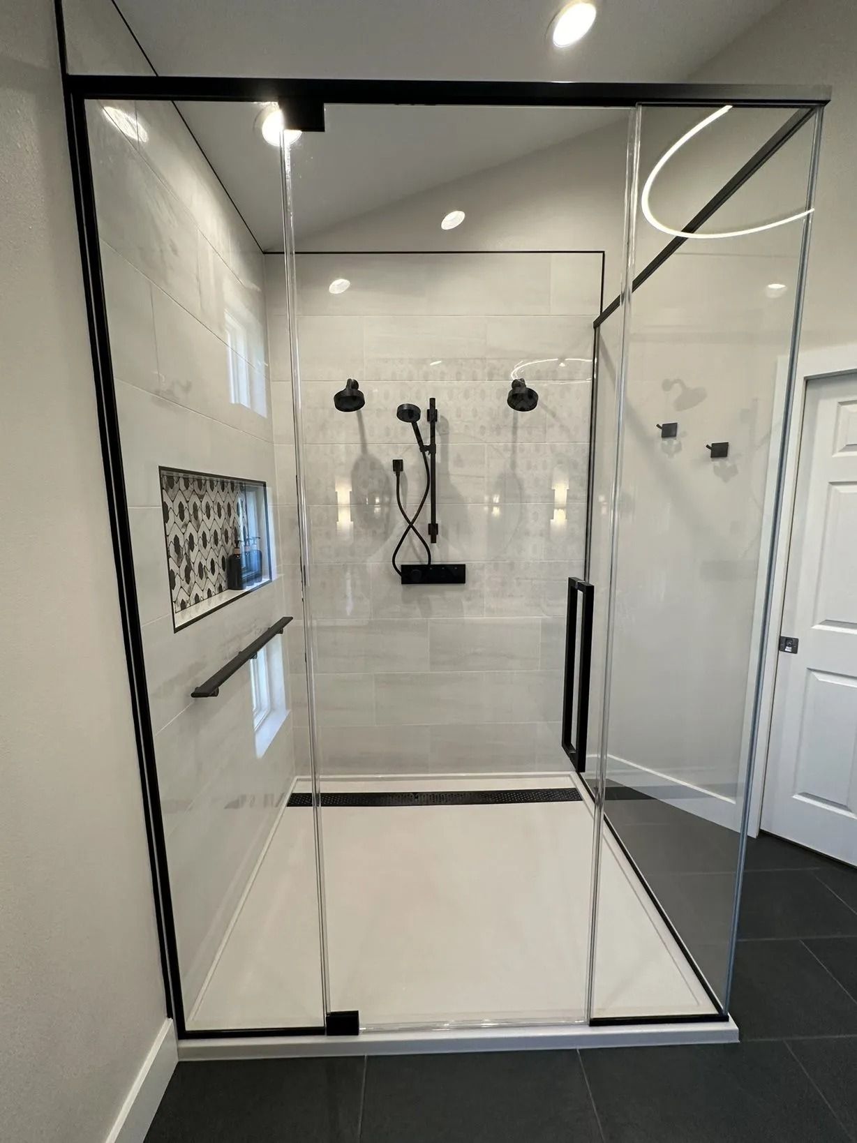 Modern shower enclosure with black trim, glass panels, and multiple shower heads on white tile.