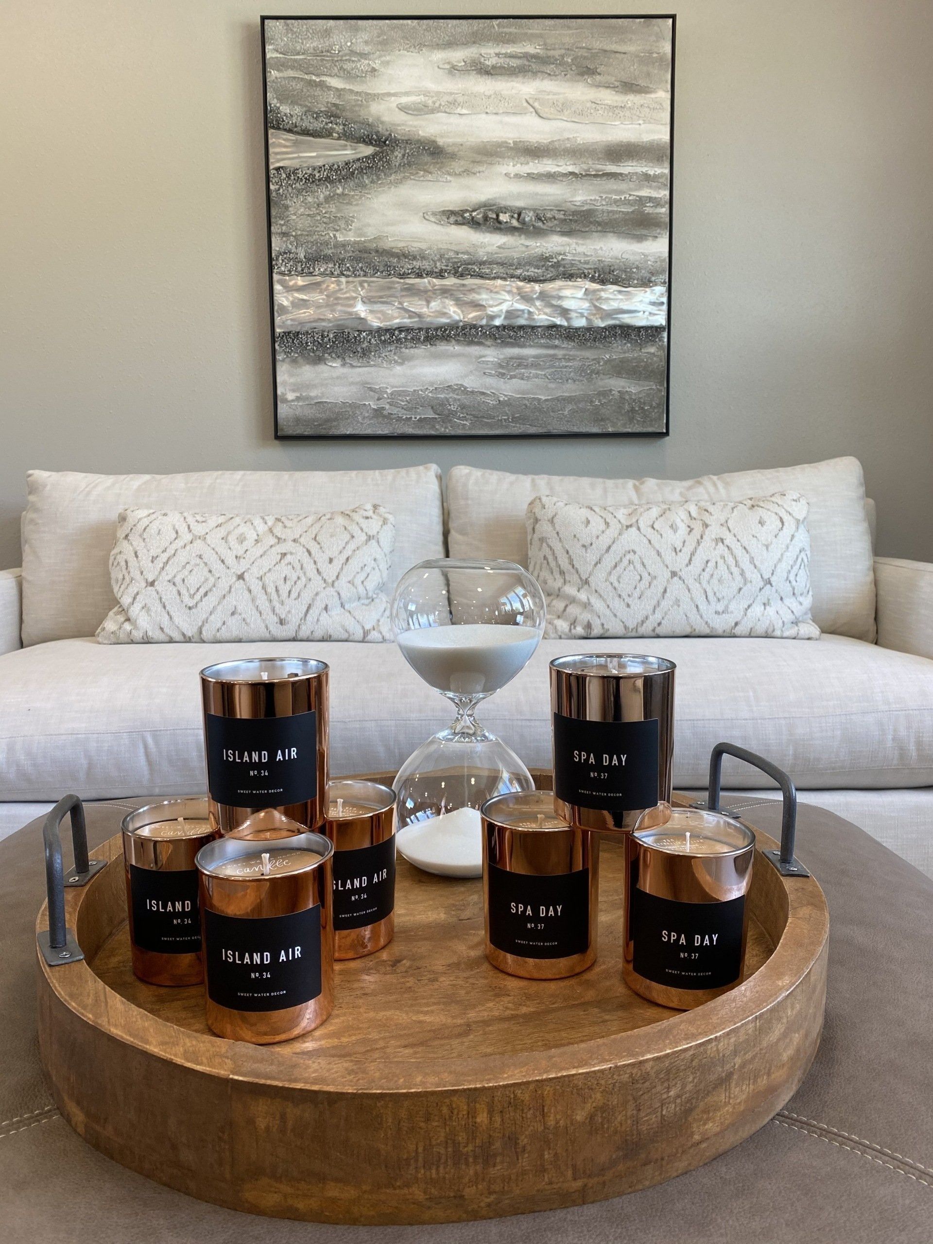 Wooden tray with candles and hourglass on a coffee table, with a sofa and artwork in the background.