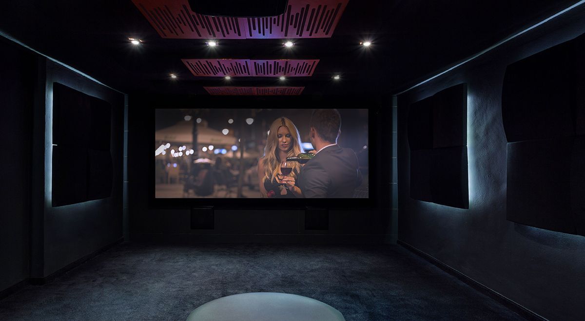A dark, modern home theater with acoustic panels, recessed lighting, and a screen displaying a movie scene.