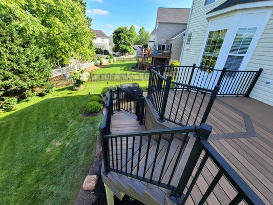 A high-angle view of a brown composite deck with black railings and stairs leading down to a green backyard lawn.