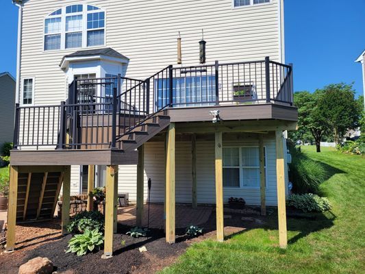 A two-story tan house features a large, elevated deck with black railings and stairs leading down to a mulched backyard.