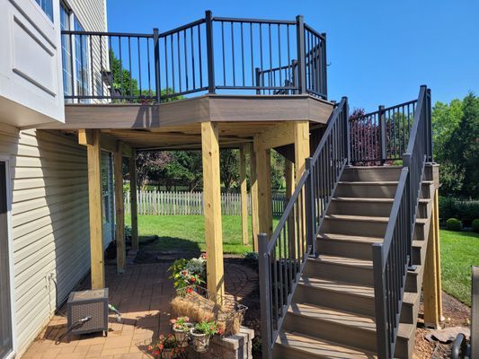 An elevated brown composite deck with black metal railings, supported by wooden posts above a paved patio with stairs.