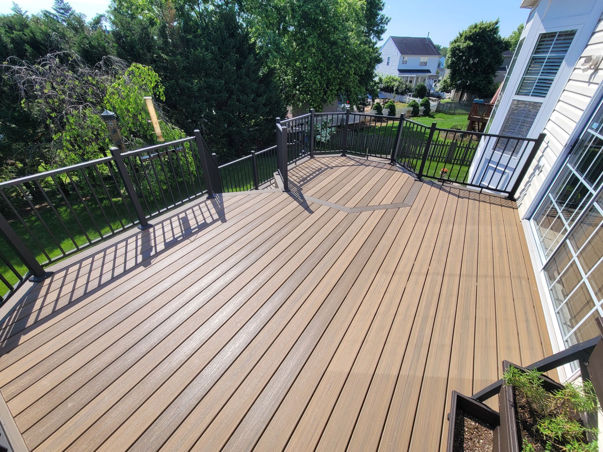 A high-angle view of a wooden deck with dark railings, extending toward a green backyard with trees and a house.