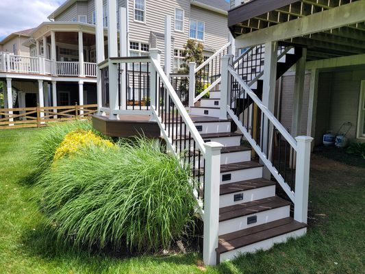 Outdoor stairs with white railings and dark wood steps lead up to a tiered deck next to a residential house.