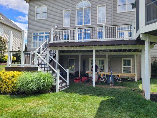 A two-story gray house with a large wooden deck, white railings, and a staircase leading down to a green lawn.