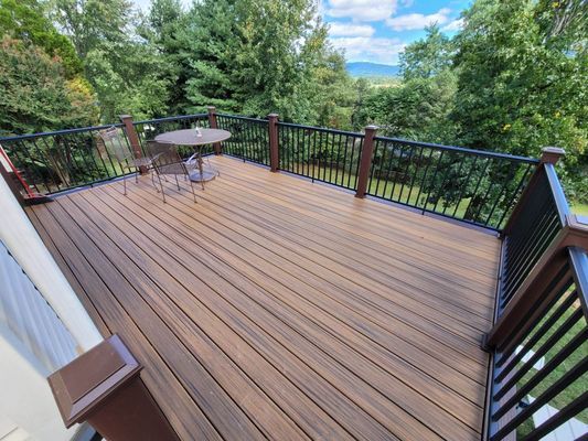 A high-angle view of a brown composite deck with black railings, a small table, and chairs overlooking a green landscape.