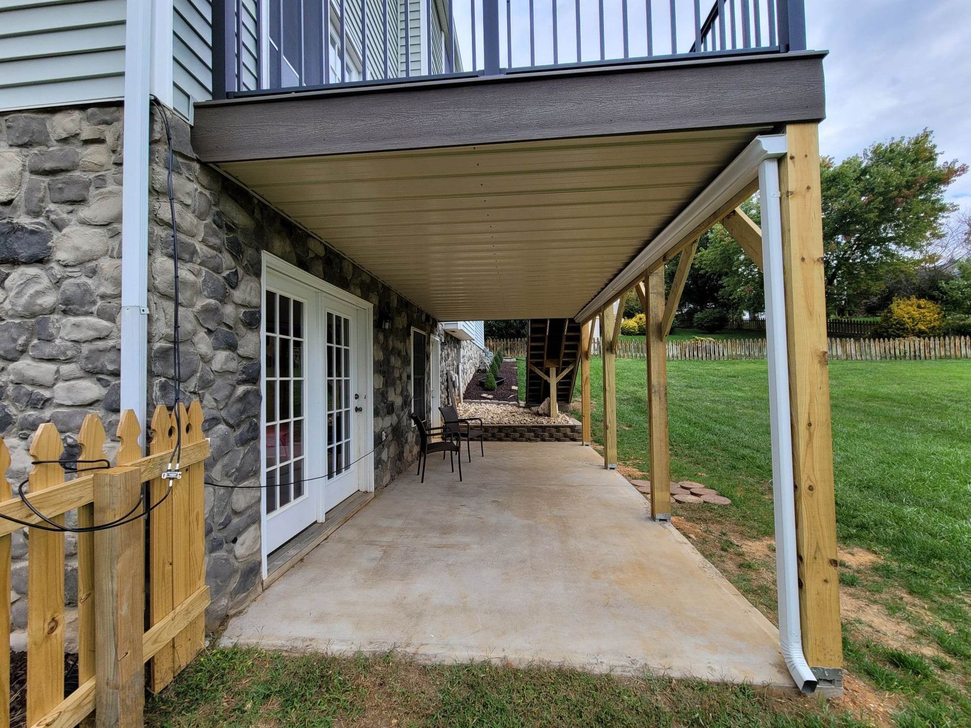A covered concrete patio under a wooden deck, next to a stone-walled house with white glass doors and a wooden fence.