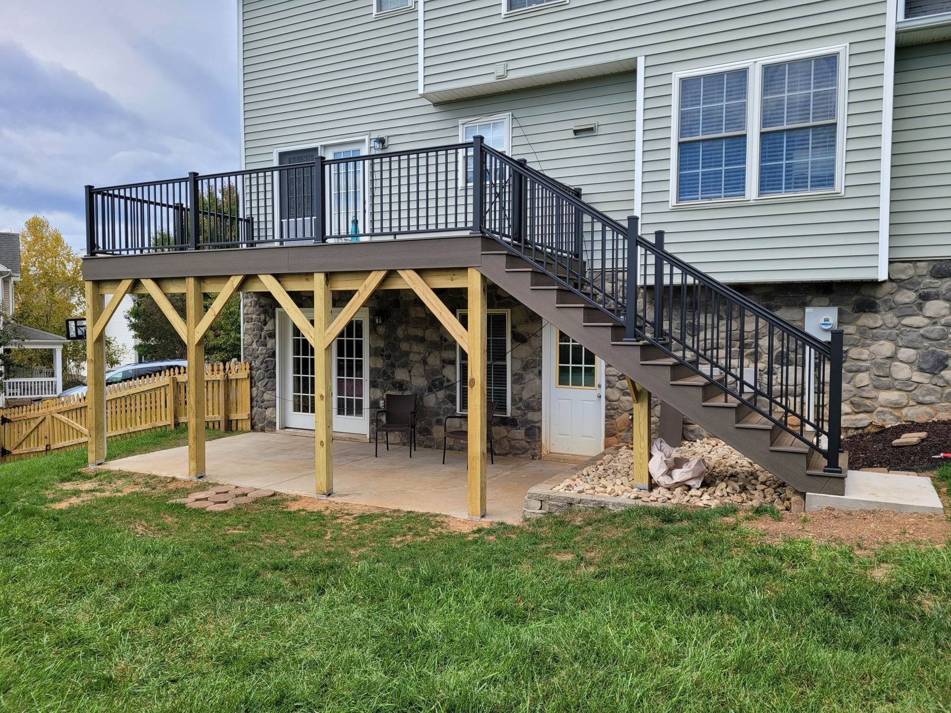 An elevated wood-frame deck with black railings and stairs over a stone-walled patio against a light green house.